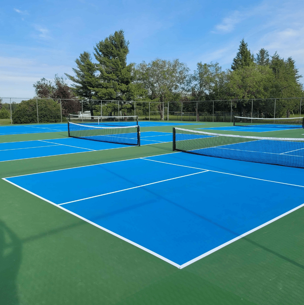 Four bright blue tennis courts are visible in a sunny outdoor setting, surrounded by trees and a fence, inviting for recreational play.