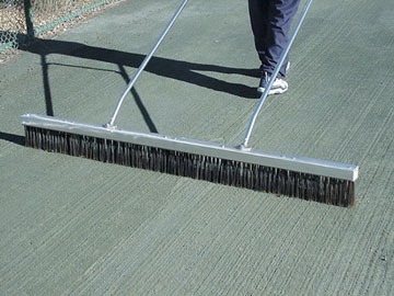 A person uses a large broom to sweep a textured green tennis court, preparing it for play by ensuring a clean surface.