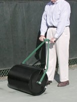 A person uses a heavy black tennis court roller, smoothing the surface to prepare it for play, ensuring optimal conditions for players.