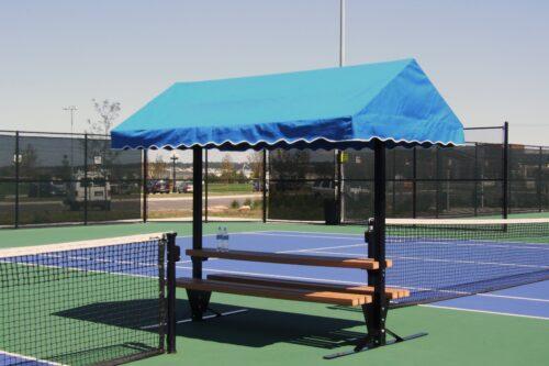 Tennis court seating area with a blue canopy providing shade over a wooden bench, ideal for players to rest between matches.