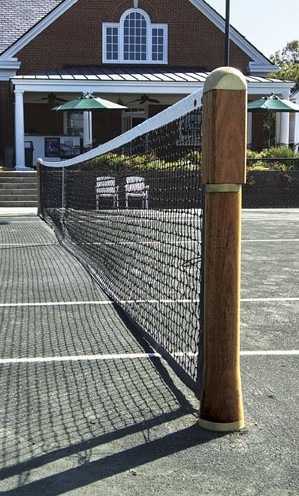 Tennis court with a wooden net post and net in the foreground; a clubhouse is visible in the background, showcasing a recreational atmosphere.