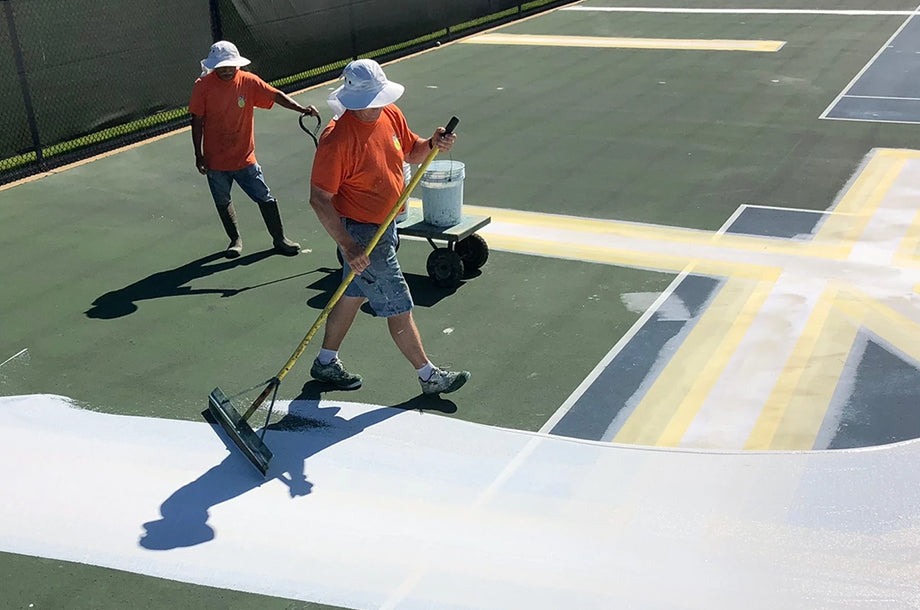 Workers in orange shirts apply white paint to a tennis court, preparing court lines and resurfacing for use. The scene highlights maintenance efforts.