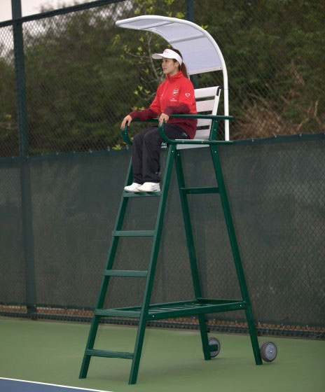 A tennis umpire sits on a tall green chair with a sunshade on a court, overseeing a match. The setting emphasizes the officiating role in the sport.