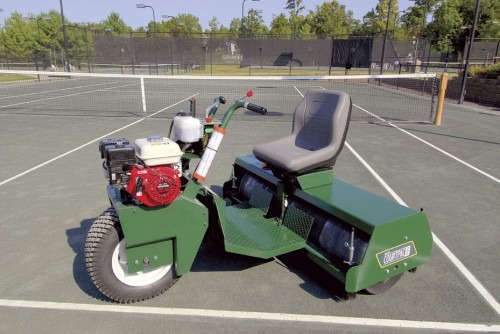 A green tennis court maintenance machine, equipped with a seat and engine, positioned on a clay tennis court, highlighting its purpose for court upkeep.