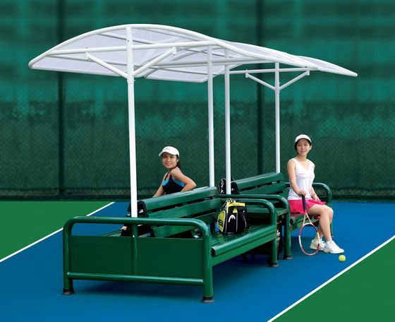 Two women sit on a green bench with a canopy on a tennis court, preparing for a game. Tennis rackets and a bag are beside them.