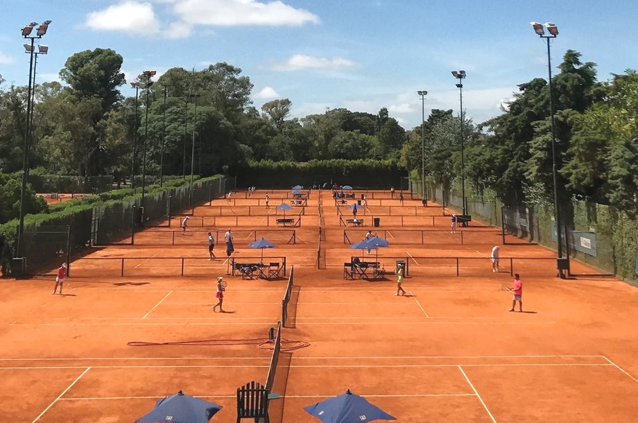 Tennis players practice on multiple clay courts under bright skies, shaded by umbrellas. The scene showcases an active sports facility.