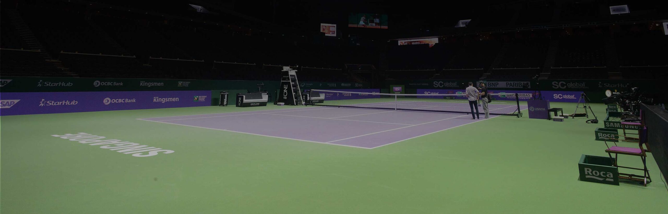 Empty indoor tennis court featuring a green and purple playing surface. Two people converse near the net, with cameras and sponsor logos visible.