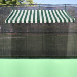 Tennis court with a green surface and a striped green-and-white awning overhead, providing shade for players and spectators.