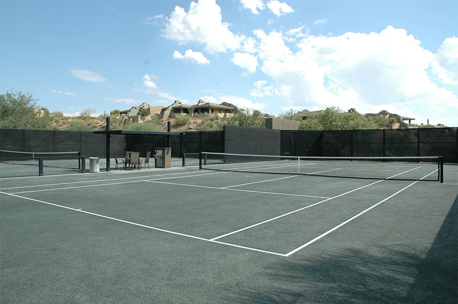 Empty outdoor tennis court with a black surface and white lines, surrounded by desert vegetation and distant hills under a partly cloudy sky.