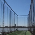 Baseball batting cage with netting, set against a clear blue sky and a river view, showcasing a training facility for aspiring players.