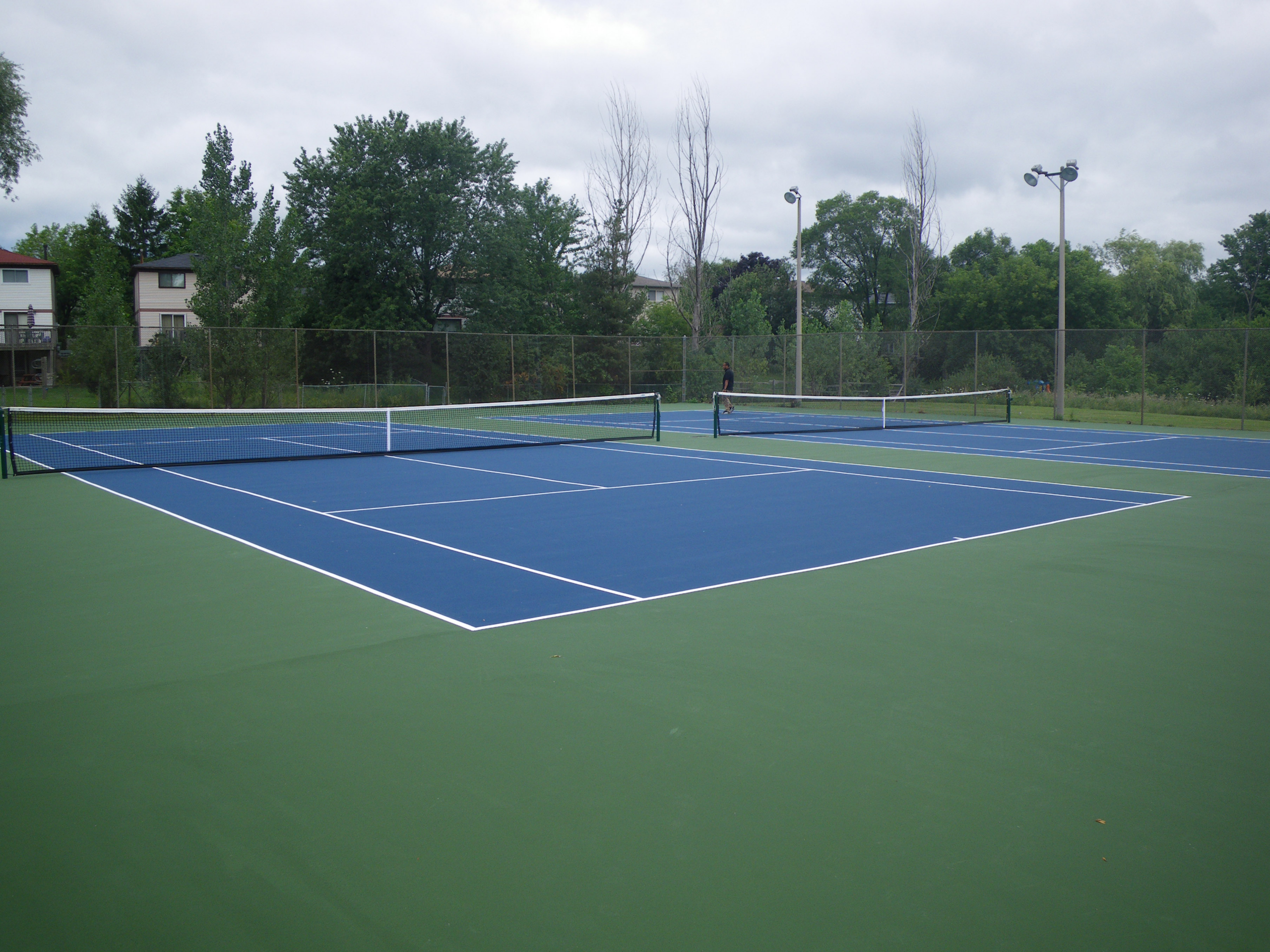 Empty tennis courts with blue and green surfaces under cloudy skies, surrounded by trees and residential buildings, ideal for recreation.