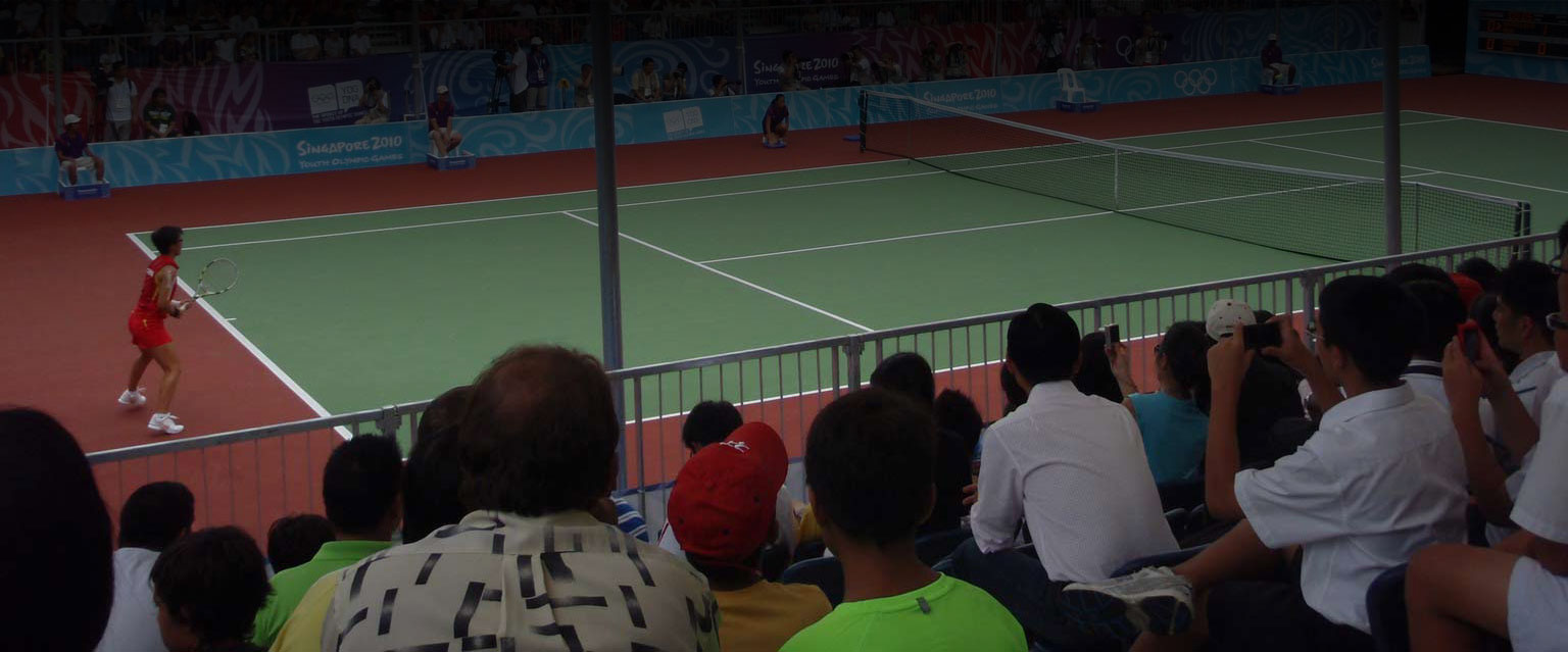 A tennis player in red prepares to hit the ball during the Youth Olympic Games in Singapore, with an audience watching eagerly in the background.