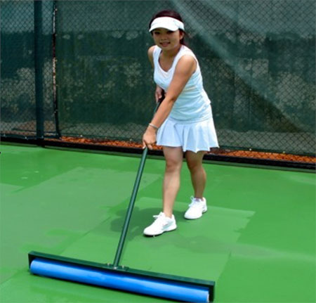 Young woman in a white tennis outfit using a roller to prepare a green court surface, ensuring it's ready for play.