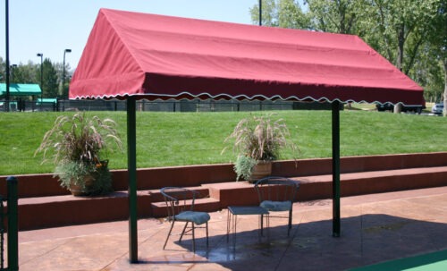 Red canopy tent with white trim provides shade over two metal chairs and potted plants on a landscaped green lawn, ideal for outdoor gatherings.