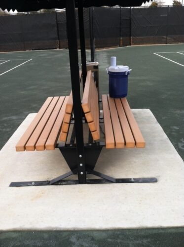 Wooden benches with a black metal frame, positioned on a tennis court, alongside a blue water cooler, providing rest for players.
