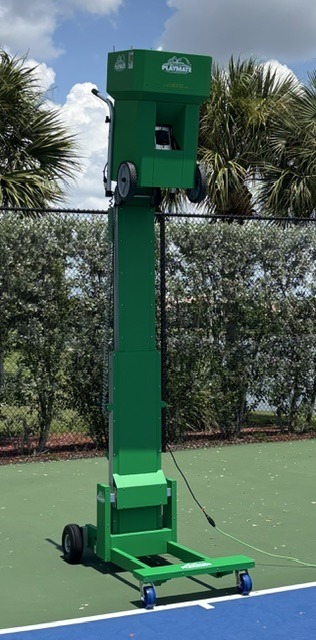 A green tennis ball retrieval system is set up on a court, featuring a tall structure with wheels for mobility, surrounded by palm trees.