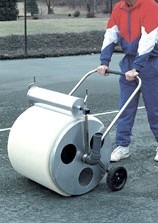 A person pushes a large, cylindrical tennis court roller across a hard court surface, demonstrating maintenance equipment for court upkeep.